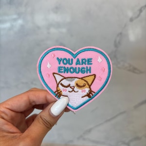 May include: A heart-shaped embroidered patch with a light pink background and a bright blue border. The text YOU ARE ENOUGH is stitched in blue above a calico cat face with closed eyes, pink cheeks, and small white star accents on the sides.