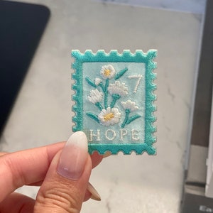 Puede incluir: Un parche bordado de color azul claro con forma de sello de correos con un diseño de margarita blanca y la palabra "HOPE" en letras blancas.