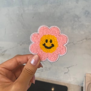 May include: A pink felt flower patch with a yellow smiley face in the centre. The flower has a white outline.