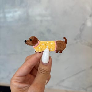 May include: Embroidered dachshund patch featuring a brown dog wearing a yellow sweater with white polka dots and pink trim. The patch is a cute, whimsical design, perfect for adding to clothing or accessories.