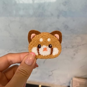 Op de afbeelding: Een bruine en witte pluche geborduurde patch van een rode panda gezicht met roze oren en neus.