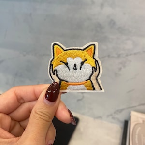 May include: Embroidered iron-on patch featuring a cartoon dog with a yellow and white face, smiling with its paws covering its face.