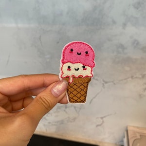 Könnte beinhalten: Gestickter Aufnäher mit einem lächelnden Eisbecher mit zwei Kugeln. Die obere Kugel ist rosa, die untere ist weiß und die Waffel ist braun. Der Aufnäher hat einen weißen Rand und ist für den Stoffaufnäher bestimmt.
