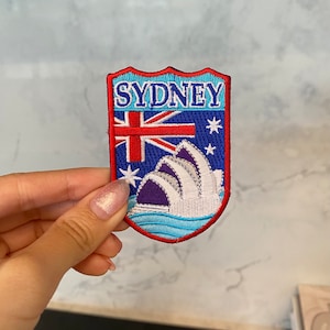 以下が含まれることがあります： 白い文字で「SYDNEY」と書かれた青と赤の刺繍パッチ。パッチにはオーストラリアの国旗とシドニーオペラハウスの画像が描かれています。