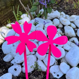 May include: Two vibrant pink flower garden stakes with a five-petal design. They are set in a garden bed with white stones and purple flowers in the background. The stakes provide a colourful accent.