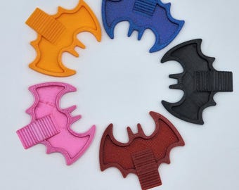 Puede incluir: Cinco pinzas para el cabello con forma de murciélago dispuestas en círculo. Cada pinza presenta un marco texturizado de color sólido en naranja, azul, negro, rojo o rosa, con una pinza tipo cocodrilo acanalada a juego en el centro.