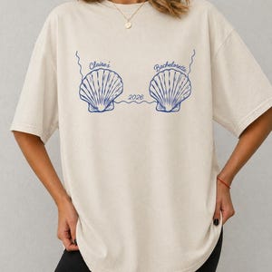 Puede incluir: Camiseta color crema con diseño de conchas marinas azules y el texto "Claire's Bachelorette 2026". La camiseta tiene un corte holgado y mangas cortas. El diseño presenta dos ilustraciones de conchas.