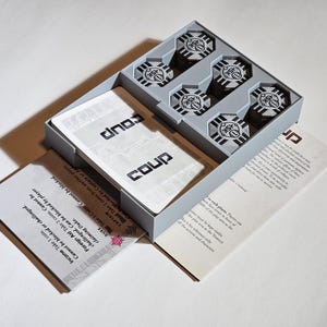 Könnte beinhalten: Eine graue Box mit einem Fach für Karten und einem separaten Fach für fünf graue Würfel mit schwarzen Markierungen. Die Box ist geöffnet und die Karten sind sichtbar. Die Karten sind weiß mit schwarzem Text, der "Coup" lautet.