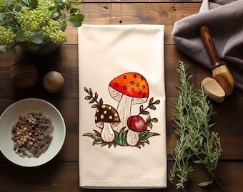 Op de afbeelding: Een witte keukendoek met een aquarel-illustratie van drie paddenstoelen. De paddenstoelen zijn rood, oranje, bruin en wit met groene stelen en bladeren.