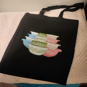 Puede incluir: Una bolsa de lona negra con un diseño colorido de una pila de tazones de mezclar vintage con patrones florales. Los tazones son en tonos de azul, verde, rosa y blanco.