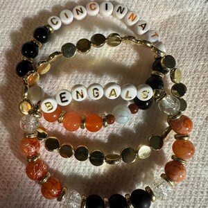 以下が含まれることがあります： 「CINCINNATI」と「BENGALS」の文字が白い文字ビーズで綴られた3つのビーズブレスレット。ブレスレットは、黒、オレンジ、金、透明のビーズを組み合わせています。ブレスレットは白い表面にあります。