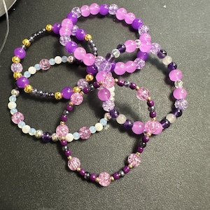 May include: A collection of beaded bracelets in shades of purple, pink, and gold. The bracelets feature various bead sizes and textures, creating a layered and colorful accessory. The bracelets are arranged in a circular pattern.