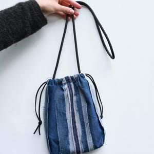 Puede incluir: Una bolsa con cordón hecha de tiras verticales de tela vaquera azul en varios tonos. El bolso presenta bordes deshilachados a lo largo de las costuras y está equipado con finas correas de cordón negro que sirven como cierre y asa.
