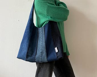 May include: A denim tote bag with a patchwork design of various shades of blue. The bag has a long strap and a rectangular shape. The bag is carried over the shoulder. The person is wearing a green sweater and black jeans.