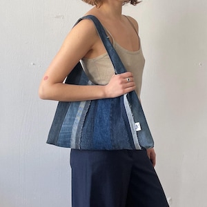 May include: A denim tote bag with a patchwork design of various shades of blue. The bag has a rectangular shape with a wide shoulder strap. The bag is held by a person wearing a neutral-colored tank top and dark blue pants.