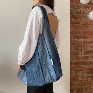 Puede incluir: Un bolso tote grande de mezclilla azul con una etiqueta blanca. El bolso está hecho de mezclilla reciclada y tiene un diseño único de patchwork.