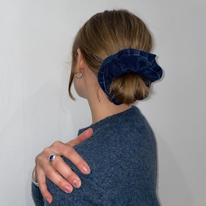 Pode incluir: Uma scrunchie de ganga azul escuro usada num coque. A scrunchie é feita de um tecido texturizado e é um tom profundo de azul. A pessoa está a usar um camisola azul e um anel de prata com uma pedra azul.