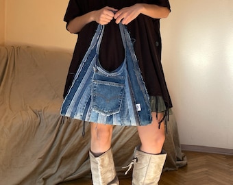 May include: A hobo-style shoulder bag constructed from repurposed blue denim fabric strips. The bag features a central patch pocket with visible stitching, frayed edges, and a triangular silhouette. The denim displays varying shades of blue.