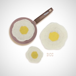 May include: A brown frying pan with a speckled interior containing a crocheted fried egg, alongside two additional crocheted fried eggs of different sizes. The eggs feature white crocheted whites and yellow centers. The text DCC is visible.
