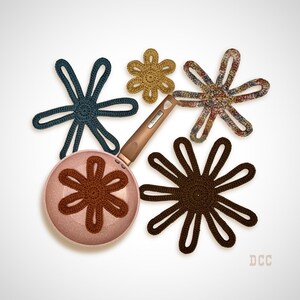 May include: A light pink speckled frying pan with a brown handle, accompanied by four crocheted flower-shaped trivets in dark blue, mustard yellow, multicolored, and dark brown. One brown crocheted flower is placed inside the frying pan.