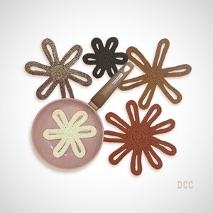May include: A pink speckled frying pan with a brown handle and five crocheted flower-shaped trivets in cream, dark brown, tan, rust, and a variegated brown-grey yarn. The trivets are arranged around the pan, with one placed inside the pan.