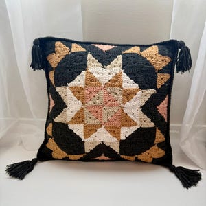 May include: Square crochet pillow with a geometric starburst pattern in black, tan, cream, and pink. The pillow has black tassels on each corner. This decorative pillow adds a touch of bohemian style to any room.
