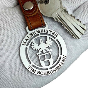 Könnte beinhalten: Ein silberner Schlüsselanhänger mit einem braunen Lederband. Der Schlüsselanhänger hat ein kreisförmiges Design mit den Worten "MALERMEISTER TIM SCHEUNEMANN" und einem stilisierten Adler und Schild.