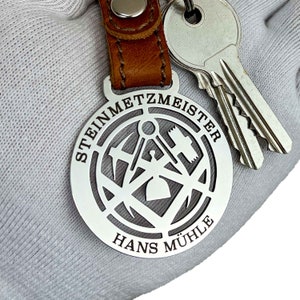 Op de afbeelding: Zilveren sleutelhanger met een bruin leren bandje. De sleutelhanger heeft een rond metalen label met een uitgesneden ontwerp van gereedschap en de tekst "STEINMETZMEISTER HANS MÜHLE".