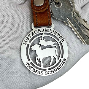 Può includere: Un portachiavi in metallo argentato con un cinturino in pelle marrone. Il portachiavi presenta un design circolare con il testo "Metzgermeister Thomas Schneider" e un'immagine stilizzata di una pecora e un pastore.