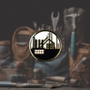 Puede incluir: Un pin de esmalte negro y dorado con una silueta blanca de un edificio de fábrica con una chimenea y ventanas.