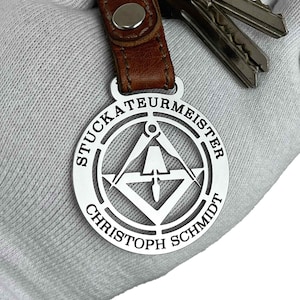 Puede incluir: Llavero de plata con una etiqueta redonda que dice "Stuckateurmeister Christoph Schmidt" y una imagen estilizada de una brújula y una escuadra.