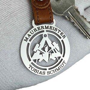 Può includere: Portachiavi in ​​argento con un cinturino in pelle. Il portachiavi presenta un design circolare con le parole "Maurermeister Tobias Schmidt" e un'immagine stilizzata di una cazzuola, un livello e un martello.