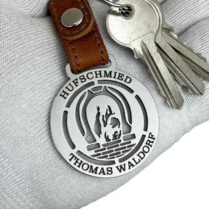 Op de afbeelding: Een zilveren sleutelhanger met een hoefijzerontwerp en de tekst "HUFSCHMIED THOMAS WALDORF".