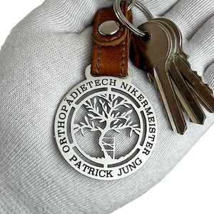 Può includere: Portachiavi in ​​argento con un cinturino in pelle e un ciondolo circolare con un design ad albero e il testo "Orthopädietechnikermeister Patrick Jung".
