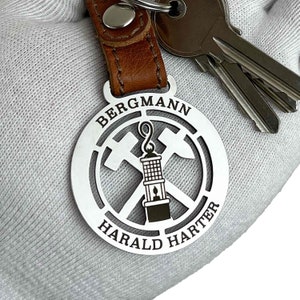 Könnte beinhalten: Silberner Schlüsselanhänger mit einem runden Metallschild, das eine Grubenlampe und gekreuzte Spitzhacken zeigt. Das Schild ist mit dem Text "Bergmann Harald Harter" graviert.