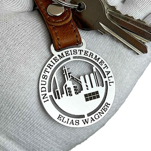 Op de afbeelding: Zilveren sleutelhanger met een uitgesneden ontwerp van een fabriek en de tekst "INDUSTRIEMEISTERMETALL ELIAS WAGNER".