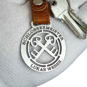 Puede incluir: Llavero de plata con una correa de cuero. El llavero presenta un diseño circular con las palabras "Schlossermeister Lukas Weber" y dos llaves cruzadas.