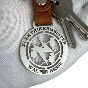 Peut inclure: Porte-clés en argent avec une sangle en cuir. Le porte-clés présente une étiquette métallique circulaire avec les mots "Elektrikermeister Walter Hesse" et un design de foudre stylisé.