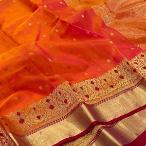 May include: A vibrant orange and red sari with gold accents. The fabric has a delicate, sheer quality, with small gold floral patterns scattered across the surface. The border features intricate gold embroidery and a red trim.
