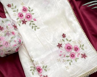 May include: A white organza saree featuring embroidered pink floral motifs with green leaves and a gold scalloped border. The fabric has a subtle sheen and a crinkled texture, complemented by a matching blouse piece with dense floral embroidery.