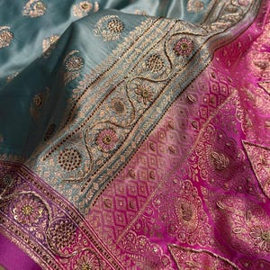 May include: Close-up of a teal and pink silk textile with intricate gold embroidery. The fabric features floral and geometric patterns, with a shiny, luxurious appearance. The design includes detailed borders and embellishments.