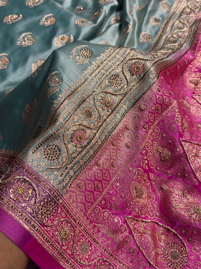May include: Close-up of a teal and pink silk textile with intricate gold embroidery. The fabric features floral and geometric patterns, with a shiny, luxurious appearance. The design includes detailed borders and embellishments.