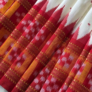 Op de afbeelding: Een textielstof met een crèmekleurig bovengedeelte met kleine rode stippen, overgaand in een levendig rood en oranje ikatpatroon. De onderste rand toont ingewikkelde geometrische motieven en horizontale accenten van gouden metaaldraad.