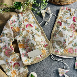 May include: White embroidered blouse with floral patterns in pink, yellow, and green. The blouse has gold trim and long sleeves. A matching bow is included. The fabric appears to be silk or a similar material.
