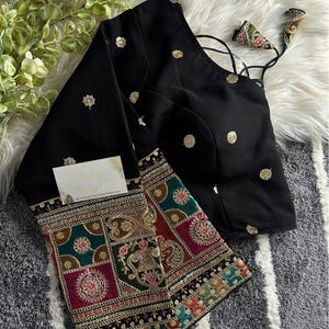 May include: Black blouse with gold detailing and elaborate embroidery on the sleeves. The sleeves showcase a colourful, geometric pattern with red, green, and gold accents. The blouse has a scoop neckline and a fitted shape.