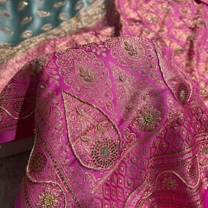 May include: Close-up of a vibrant pink textile with intricate gold embroidery. The design features paisley shapes, floral patterns, and geometric borders. The fabric has a luxurious sheen, suggesting a high-quality material.