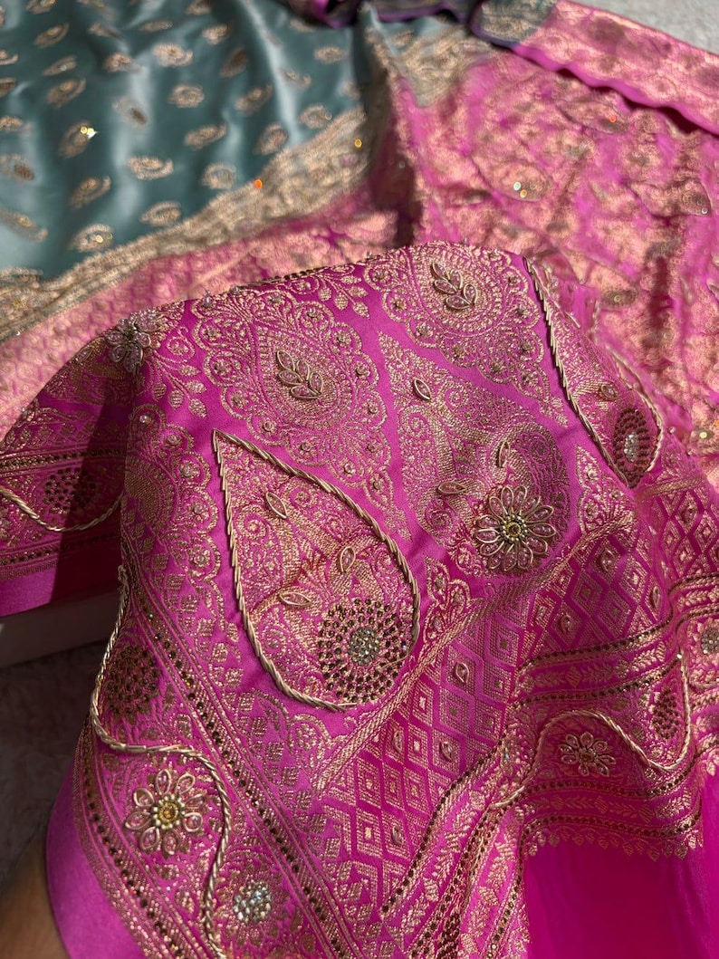 May include: Close-up of a vibrant pink textile with intricate gold embroidery. The design features paisley shapes, floral patterns, and geometric borders. The fabric has a luxurious sheen, suggesting a high-quality material.