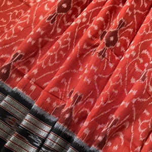 May include: A woven fabric featuring a vibrant red base with a repeating white and dark brown floral ikat pattern. The bottom edge is finished with a wide black border containing intricate silver and white geometric diamond motifs and stripes.