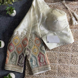 Pode incluir: Blusa marfim de um ombro com bordados intrincados nas mangas. As mangas apresentam um design colorido, semelhante ao paisley, com tons de amarelo, laranja, rosa, verde e azul. A blusa tem um corpete ajustado e enfeites delicados.