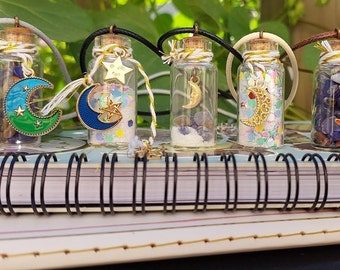May include: Five glass bottle necklaces with cork stoppers and metal charms. Each bottle contains different colored glitter, crystals, and a crescent moon charm. The necklaces are displayed on a spiral bound notebook.
